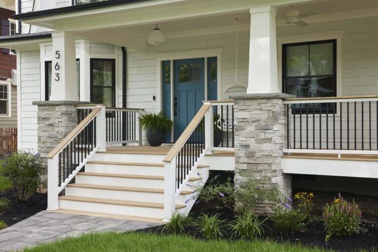 A covered front porch showcases a stark blue door with lush green ferns in attractive planters on either side. A macramé glider adds even more personality to the porch. Two stone columns lead up to tapered white PVC column wraps on either side of the stairs. The steps are made of TimberTech AZEK Weathered Teak boards from the Vintage Collection and provide gorgeous accents to the already personality-filled porch.